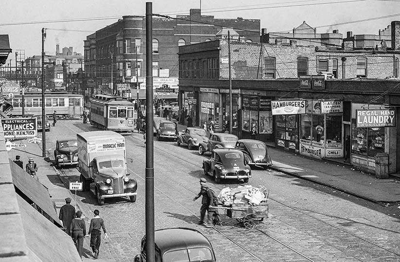 Black and white historical street photograph showing vintage cars, buses, pedestrians, storefronts, and utility poles in an urban setting.