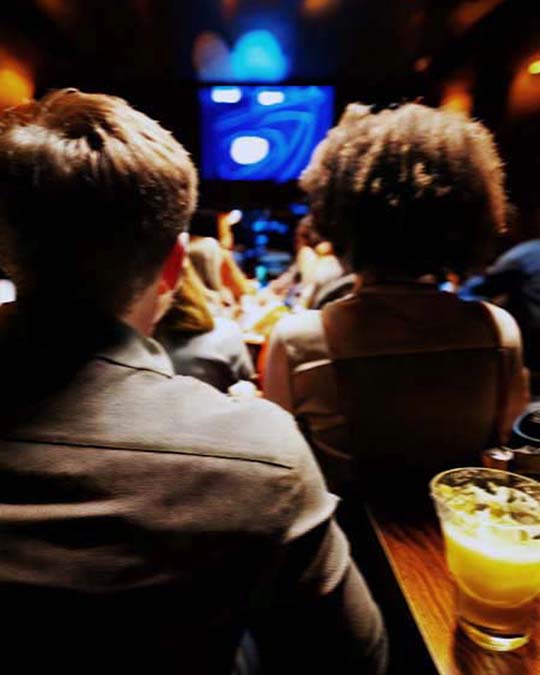 A dimly lit bar or lounge scene viewed from behind two seated people, with a glowing blue screen in the background and an orange drink on a table in the foreground.