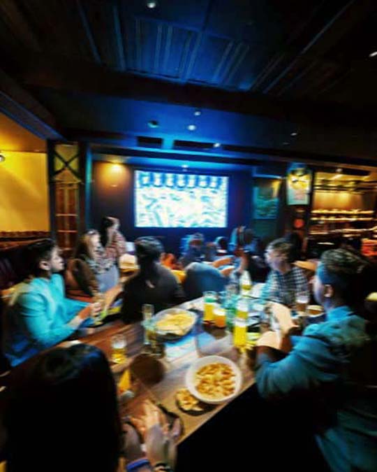 A group of people gathered around a bar table in a dimly lit restaurant or bar, watching a large projection screen on the wall. The scene is illuminated by warm yellow ambient lighting and blue overhead lights, with plates of food and drinks on the table.