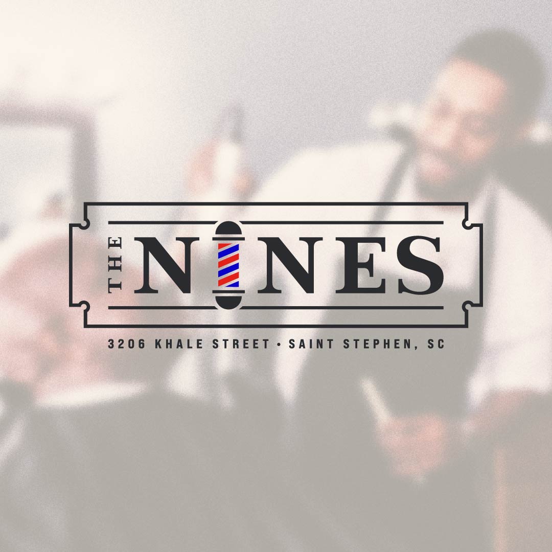 A square graphic with a blurred background photo of a barber shop interior and a barber at a mirror. Centered over the photo is a ticket-style logo reading “THE NINES,” with a barber pole graphic replacing the letter N.