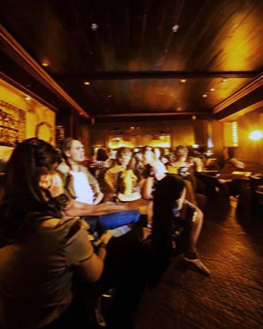 A dimly lit indoor event space or club with several people seated and facing a stage or screen, illuminated by warm yellow overhead lighting.
