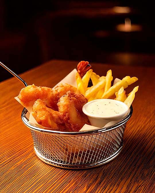 A metal wire basket filled with golden fried food, including a piece of fried fish, french fries, and a small white dipping sauce cup, set on a wooden table.