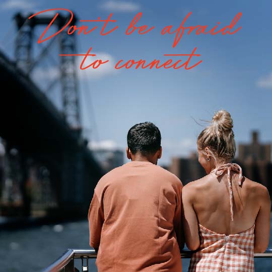 A couple seen from behind standing by a railing and looking out at a city waterfront with a bridge and blue sky. Large red script text across the top reads, 'Don't be afraid to connect.