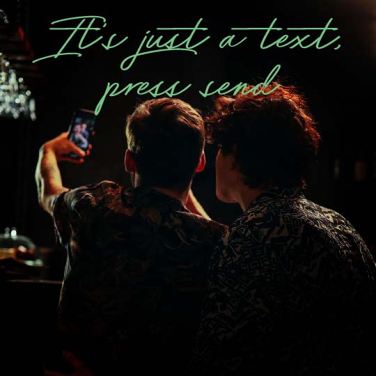 Two people seen from behind in a dimly lit bar or cafe setting, with one holding up a smartphone to take a selfie. Large neon-style green text across the top reads, 'It's just a text, press send.