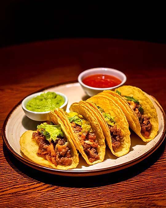 Four soft corn tacos filled with seasoned meat and topped with chopped green onions, served on a beige ceramic plate with two small dipping bowls of salsa and guacamole on a dark wooden table.
