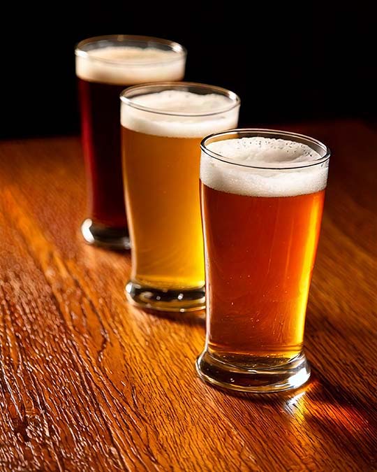Three glasses of beer arranged diagonally on a wooden bar table, with the front glass showing an amber beer and white foam head, and the back glasses showing darker and lighter beer tones against a dark background.