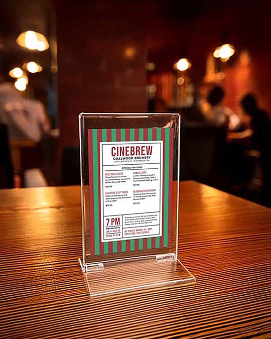 A clear acrylic tabletop menu holder displaying a 'Cinebrew Collegiate Brewery' menu, set on a wooden table with a blurred restaurant background.